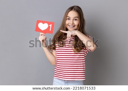 Portrait of adorable smiling little girl wearing striped T-shirt pointing at heart like icon, recommending to click on social media button. Indoor studio shot isolated on gray background.