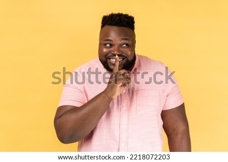 Portrait of optimistic positive man wearing pink shirt standing, showing hush sign and looking away with toothy smile, sharing secret. Indoor studio shot isolated on yellow background.