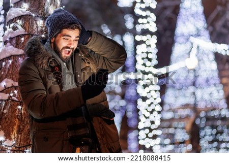 Cheerful handsome young man having video call using smart phone while celebrating New Year in the city streets with Christmas lights all around