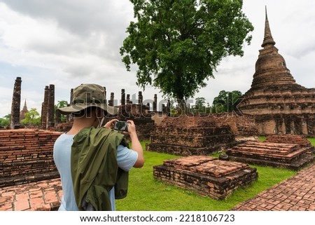 Asian man wearing green hat is using a mobile phone. He stood to take pictures of ancient monuments, pagodas, viharn, mondop, and church in the Sukhothai Historical Park. Sukhothai Province, Thailan