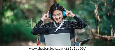 Happily Young woman sitting on camping chair wearing headphone listening music from laptop computer while relax on camping in forest