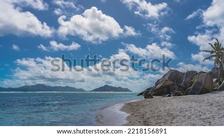 Picturesque granite cliffs rise on a tropical beach. The waves of the turquoise ocean spread over the sand. In the distance, silhouettes of islands are visible against the blue sky and clouds. 