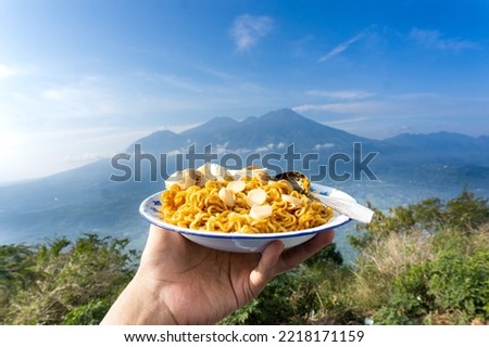 A man's hand holding a plate of instant noodles, with mountain as background. Outdoor or camping photo concept.