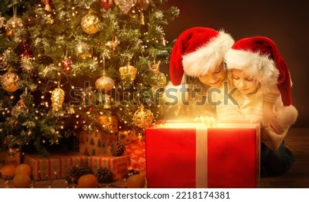 Christmas Children opening shining Red Present. Kids in Santa Hat looking at Light inside Gift Box next to decorated Xmas Tree in Dark Home Room. Christmas Family Sisters