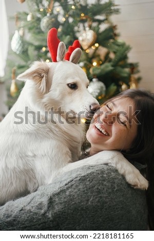 Merry Christmas! Cute dog in reindeer antlers playing with owner at stylish christmas tree. Pet and winter holidays. Happy woman hugging adorable funny white danish spitz in festive room