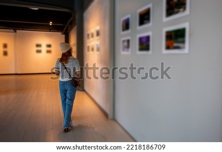 Asian woman walking through art gallery collection in front framed paintings pictures on white wall, Visitor person walk looking to picture at photo frame at interior show exhibition artwork gallery Royalty-Free Stock Photo #2218186709
