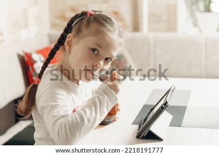A little girl eats something from a glass cup and watches cartoons on a tablet while sitting at a table in the kitchen.