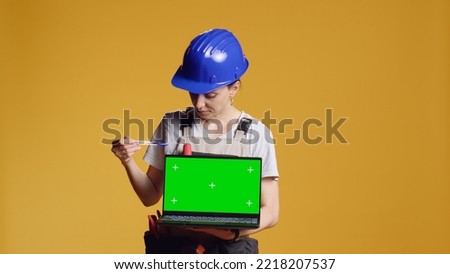 Portrait of handywoman holding laptop with greenscreen display over yellow background, using blank chroma key template with isolated mockup copyspace. Working on renovation with computer.