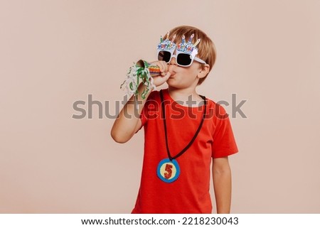 Excited amazing joyful little boy wearing red shirt and party sunglasses. Funny Little children boy plays paper trumpet, celebrates birthday. Studio shot isolated over beige background. Party concept.