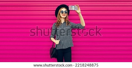 Portrait of happy smiling young woman taking selfie with smartphone wearing black round hat on pink background
