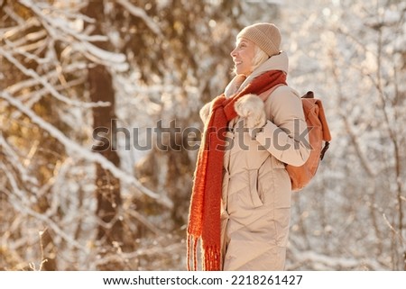Side view portrait of smiling senior woman enjoying hike in winter forest lit by sunlight, copy space