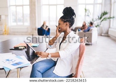 Side view of smiling African American female freelancer in casual clothes with digital tablet sitting at wooden table while working on project in office