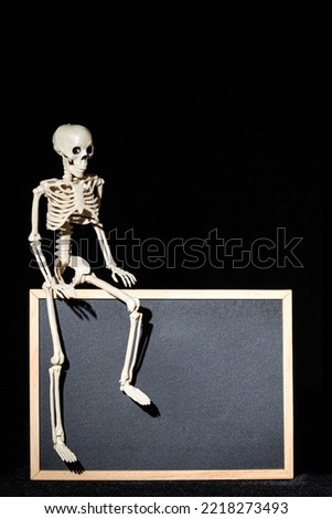 Funny human skeleton sitting on an empty black flipchart on a black background, copy space, mockup.