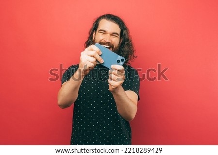 Photo of joyful young bearded hispter man with long hair playing online games at his smartphone over pink background.