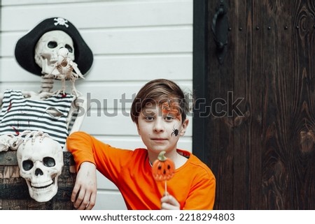 Young teen boy with painted face spider at all hallows eve photo session enjoy lollipop pumpkin Jack indoor. Stylish child in orange clothes celebrates halloween holiday of human skeleton pirate skull