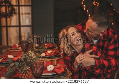 Portrait of young lovely couple hugging indoor eve 25 December. Lovers laughing hugs kisses waiting miracle at home. Celebrating new year garlands lights noel in red plaid pajamas passion tenderness