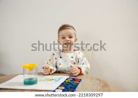 An adorable male toddler dressed in colorful dots sits at a table and draws with a brush and watercolors on paper. He is happy as he draws and has a warm expression and a sweet smile Looking at camera