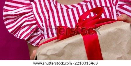 The child is holding a large gift wrapped in kraft paper with a red ribbon. For Christmas, New Year, Valentine's Day, March 8, Mother's Day. High quality photo