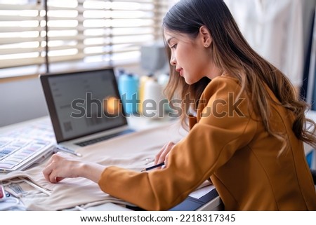 Young professional clothes fashion designer sitting near sewing machine use laptop computer and tablet pc to reference and draw the design on the paper pattern