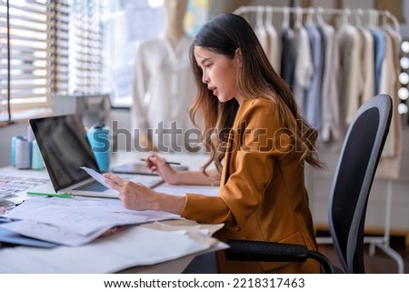 Young professional clothes fashion designer sitting near sewing machine use laptop computer and tablet pc to reference and draw the design on the paper pattern