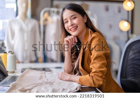 Young professional clothes fashion designer sitting near sewing machine use laptop computer and tablet pc to reference design smiling portrait to camera