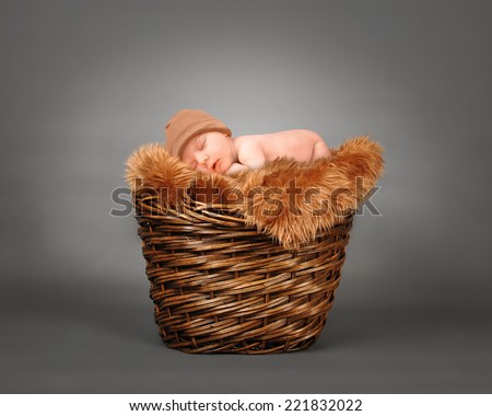 A cute little baby is sleeping in a wooden basket with brown fur and is wearing a hat. The baby could be a boy or girl on a isolated gray photography backdrop for a parenting or love concept. 