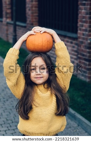 Funny child girl in orange pullover for Halloween with pumpkin and on a dark brick background. Happy laughing child girl in orange pullover and black jeans to halloween.