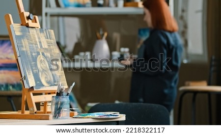 Abstract painting black canvas with gold drawings on easel stands on table in workshop studio with paint brushes red-haired unrecognizable woman artist painter choose acrylic oil preparing for drawing