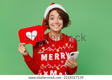 Merry young woman wears xmas sweater Santa hat posing hold in hand use mobile cell phone heart form like icon sign isolated on plain pastel light green background. Happy New Year 2023 holiday concept