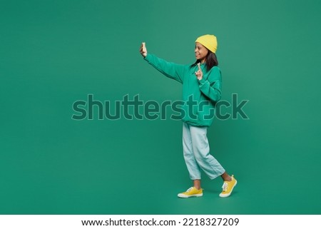 Full body little kid teen girl of African American ethnicity 13-14 year old wear casual hoody hat do selfie shot on mobile cell phone show v-sign isolated on plain green background Childhood concept