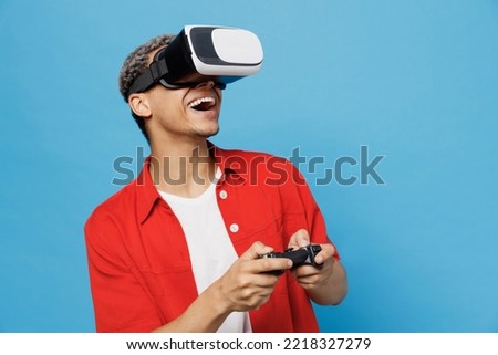 Young amazed happy man of African American ethnicity wear red shirt hold in hand play pc game with joystick console watching in vr headset pc gadget isolated on plain pastel light blue cyan background