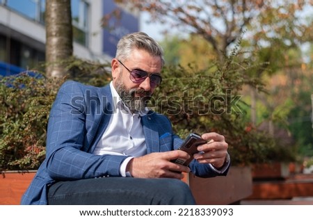 A handsome senior business man using mobile phone app texting while sitting on the park bench.