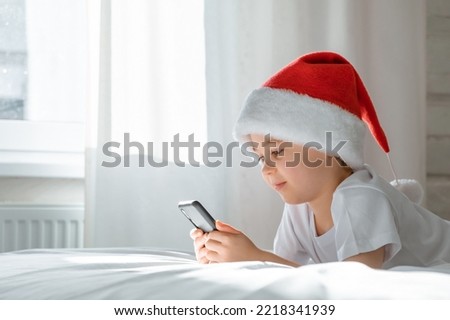 5 year smiling boy is lying on bed looking at the screen and playing smartphone game. Close-up portrait. Holds cell phone. Online education. White clothes. Telephone user. Save child vision concept.