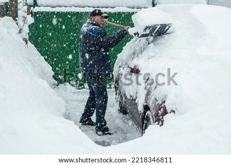 A man in a cap is shoveling snow off a car that was covered in snow during the snowfall. Large flakes of snow are falling. A snowy winter in Europe. The global of climate change Royalty-Free Stock Photo #2218346811