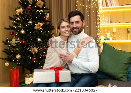 Portrait of mature family bearded man and blonde woman looking at camera hugging near Christmas tree, couple celebrating christmas and new year with gifts at home sitting on sofa in living room.