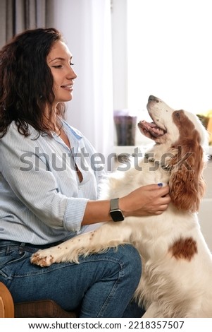 Cheerful young woman holding her big puppy with black nose and laughing. Indoor portrait of smiling girl with dark short hair posing with dog on background at home.