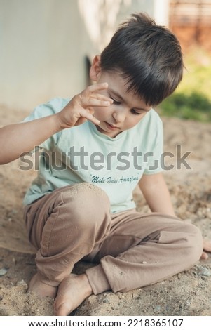 Portrait of lonely kid sitting alone on sand in the garden, looking down deep in though. Nature summer. Warm sunny day. Active outdoors games for kids in summer