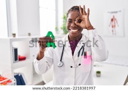 Beautiful black doctor woman holding support green ribbon for mental health awareness smiling happy doing ok sign with hand on eye looking through fingers 