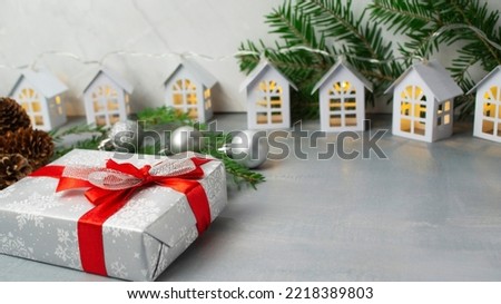 The gift is packed in gray paper with a red bow. In the background, garlands and branches of spruce with silver balls. Flat lay on a gray background.