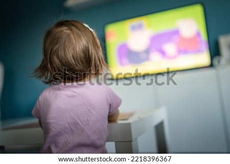 Back view of one unknown caucasian child toddler girl standing at home watching tv while eating copy space childhood growing up development leisure concept