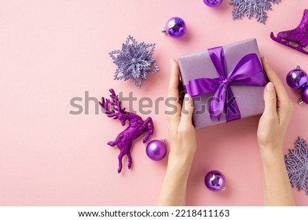 Christmas concept. First person top view photo of girl's hands holding violet giftbox over lilac baubles snowflake flower ice skates deer ornaments on isolated light pink background with copyspace