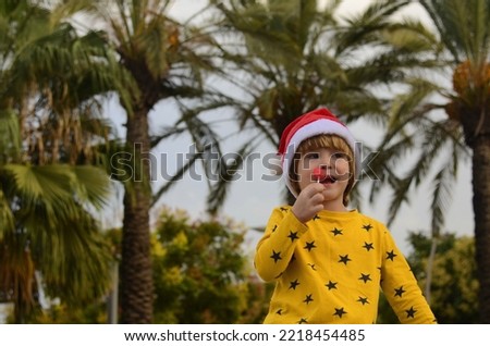 Portrait of a positive kid in a santa claus hat under a palm tree. New Year and Christmas in a tropical island, winter holidays, exotic holiday. Place for text. Boy's face close up.