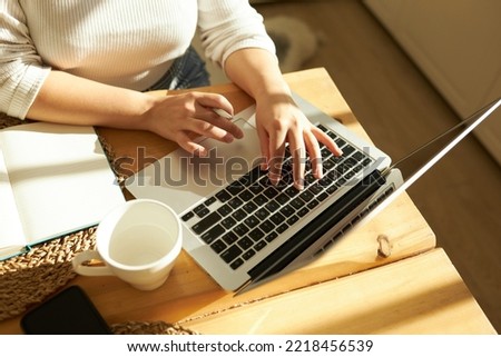 Cropped shot top view of female hands typing on keyboard of opened laptop at kitchen table with white coffee mug and notebook, studying online, checking e-mail, using learning application