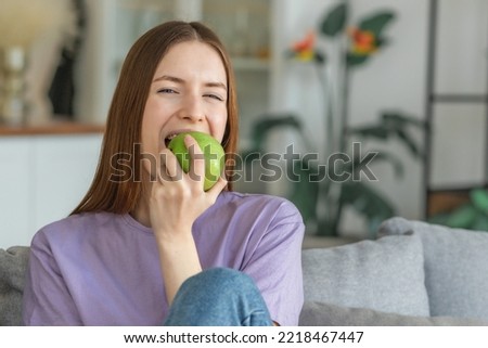 Healthy lifestyle concept. Beautiful young Caucasian woman biting fresh green apple sitting on the couch at home, looking at the camera Royalty-Free Stock Photo #2218467447
