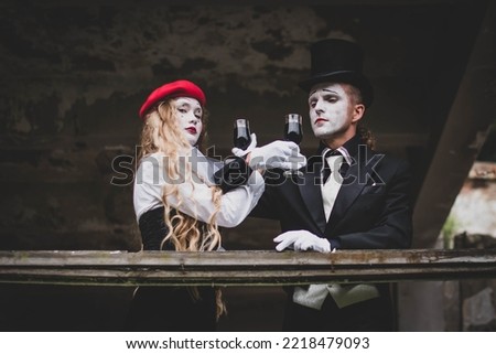 The hatter and alice in wonderland. A girl with long hair in a red hat with white makeup on her face. A man in a suit with make-up and a black hat. They drink wine. Halloween