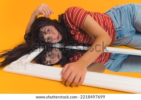 Teenagers with psychological problems such as body dysmorphia. Sad long-haired female caucasian teenager in stripped t-shirt lying next to bedroom mirror and touching in with her hand. Studio shot Royalty-Free Stock Photo #2218497699
