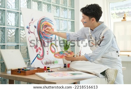 Young man holding paint brushes and palette, painting on canvas at studio.
