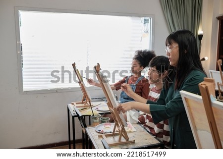 A female Asian teacher teaches and demonstrates to the children on acrylic color picture painting on canvas in art classroom, creatively learning with skill at the elementary school studio education.