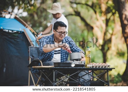 Portrait of Asian traveler man glasses frying a tasty fried egg in a hot pan at the campsite. Outdoor cooking, traveling, camping, lifestyle concept. Royalty-Free Stock Photo #2218541805