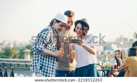 Pretty young woman is using smartphone with her cheerful male friends, young people are laughing and watching funny photos.